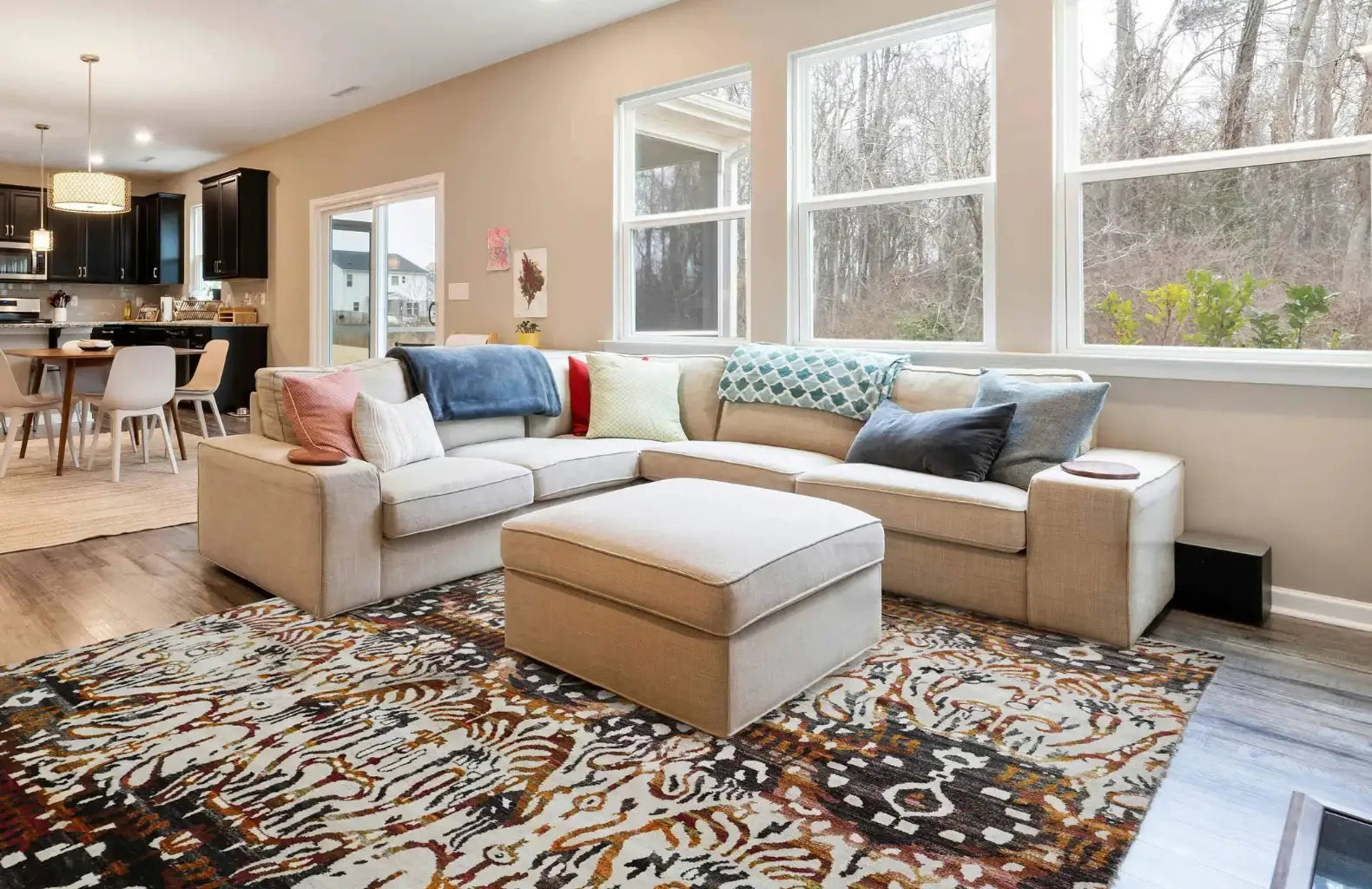 Spacious living room with a beige sectional sofa adorned with colorful pillows, a patterned rug, large windows with a forest view, and an adjoining kitchen with dark cabinets. The atmosphere is cozy and inviting.